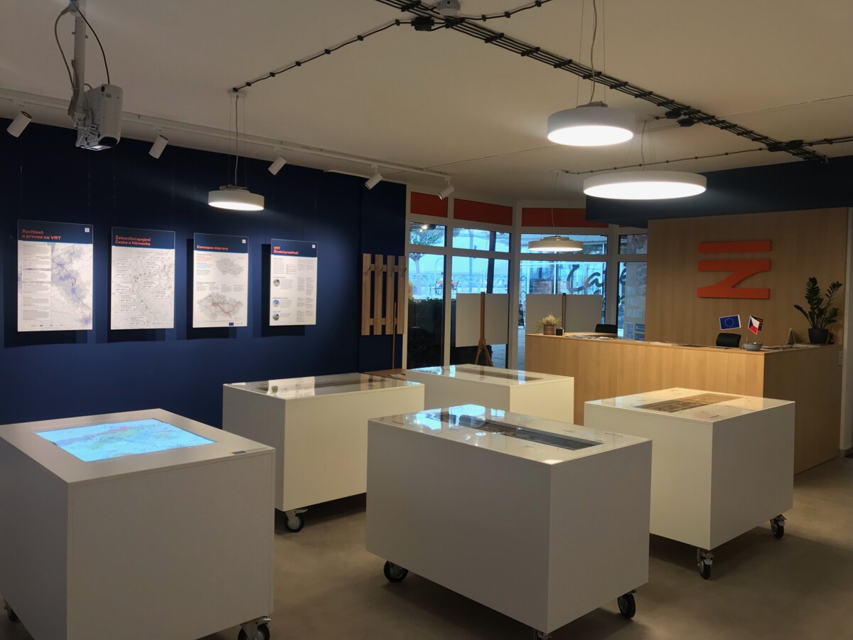 Inside the information centre in Ústí nad Labem, the dark blue walls are decorated with information boards and the lighting is atmospheric. The orange logo of správa železnic adds a vibrant touch. Positioned in the middle of the room are white tables on wheels.