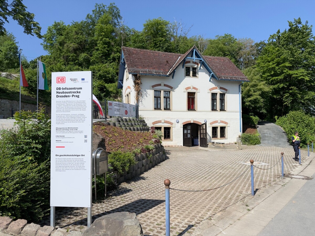 The entrance door of the white building of the information centre in Heidenau is open. Behind the building, there are numerous trees, and it is a sunny spring day. On the left side of the picture, in front of the building's entrance, there is a Deutsche Bahn information board.
