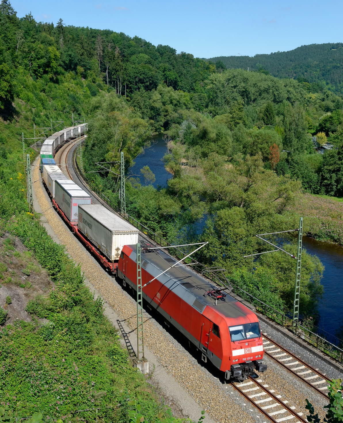 On a clear day, a lengthy red freight train, travels through a lush green landscape. The scenery consists of forested areas along the edges and a blue river.
