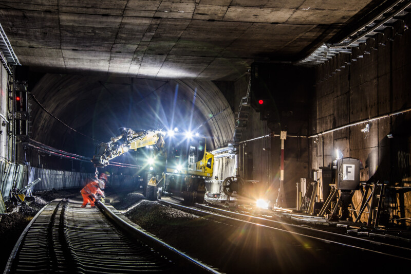 A construction site in a tunnel at night: The bright lights illuminate large pieces of construction equipment and create harsh contrasts with their shadows. A worker wearing a high-visibility waistcoat can be seen next to a railway track. 